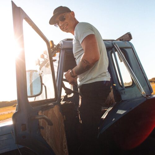 A man in sunglasses stands by a blue tractor in sunlit countryside, exuding casual confidence.